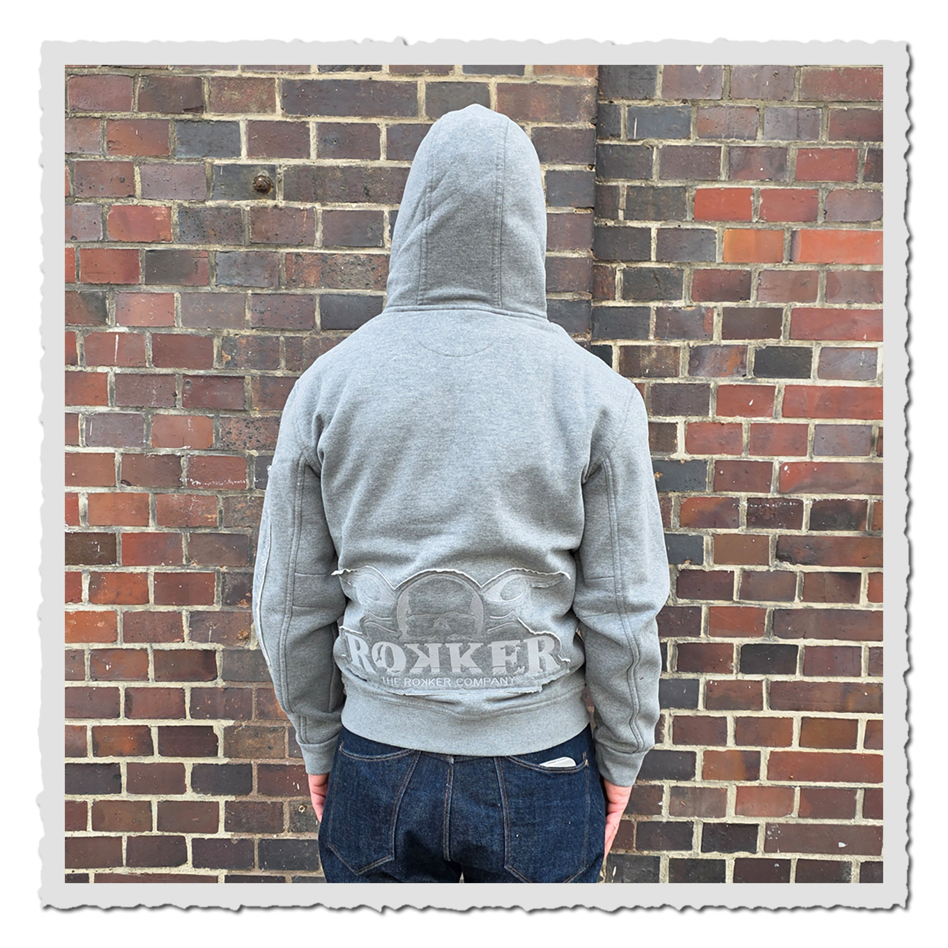 Person in hellgrauem Zip Hoodie von hinten vor Backsteinwand, Kapuze aufgesetzt, dunkle Jeans sichtbar.